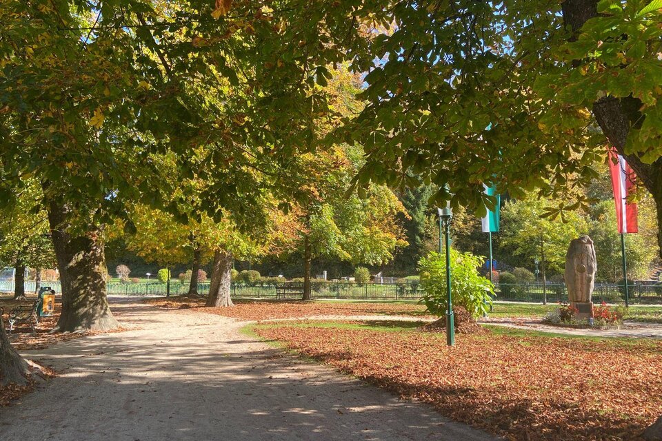 <p>Der Kurpark im Zentrum von Bad Aussee im Herbst. Ein Weg führt durch den Park. Welke Blätter liegen am Weg und auf der Wiese. Auf der rechten Seite steht der große Mittelpunkt Österreichs Stein mit zwei gehissten Flaggen, der steirischen und der österreichischen Flagge.</p> | © Kurpark, Bad Aussee (c) TVB Ausseerland Salzkammergut