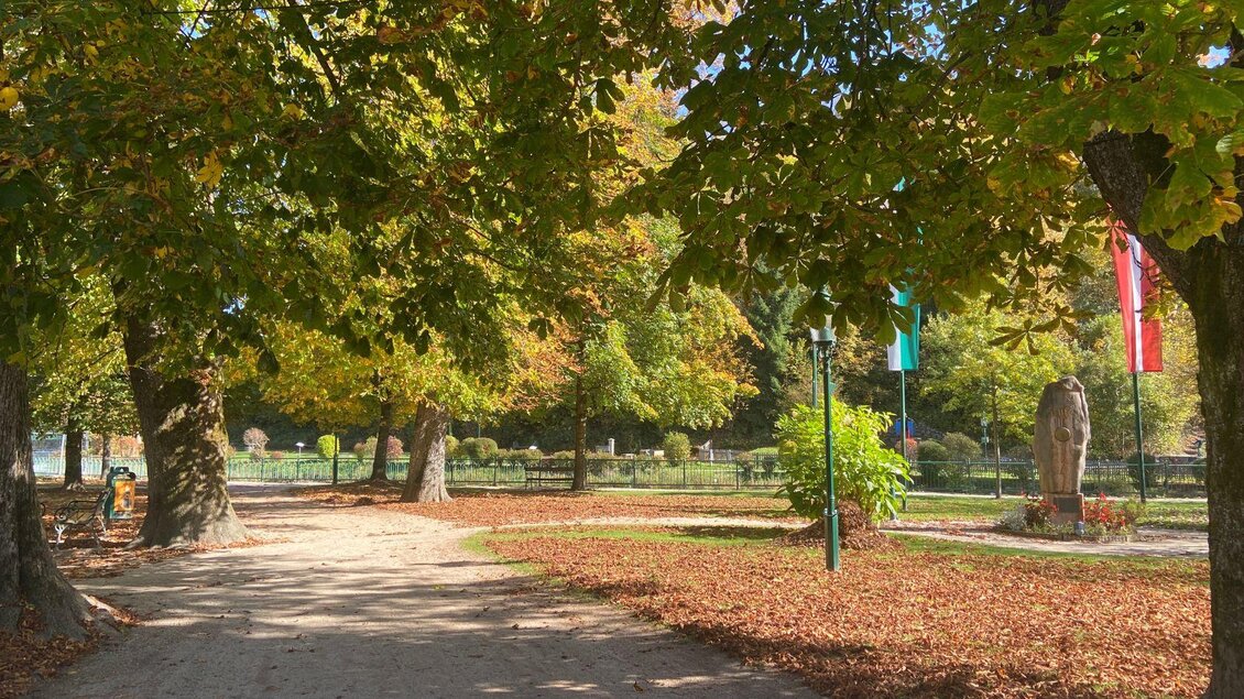 <p>Der Kurpark im Zentrum von Bad Aussee im Herbst. Ein Weg führt durch den Park. Welke Blätter liegen am Weg und auf der Wiese. Auf der rechten Seite steht der große Mittelpunkt Österreichs Stein mit zwei gehissten Flaggen, der steirischen und der österreichischen Flagge.</p> | © Kurpark, Bad Aussee (c) TVB Ausseerland Salzkammergut