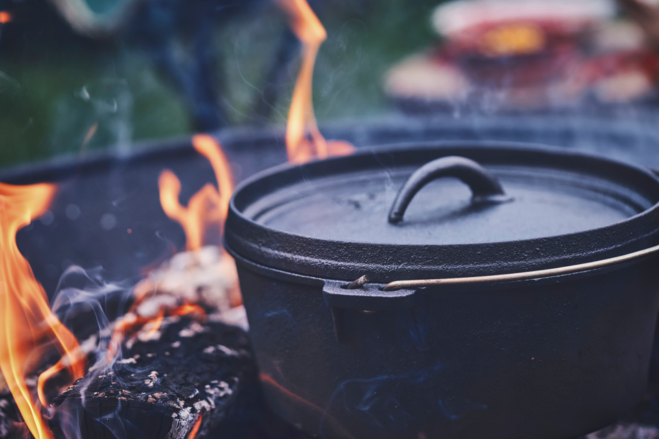 Ein schwarzer Kochtopf steht über einem offenen Feuer. Flammen leuchten hell und erzeugen eine gemütliche Atmosphäre im Freien. | © Canva