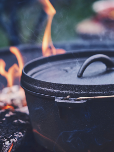 Ein schwarzer Kochtopf steht über einem offenen Feuer. Flammen leuchten hell und erzeugen eine gemütliche Atmosphäre im Freien. | © Canva