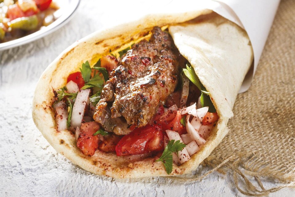 Ein frisch zubereitetes Wrap mit saftigem Fleisch, Tomaten, Zwiebeln und Kräutern. Daneben sieht man eine kleine Schale mit weiteren Beilagen. | © Canva