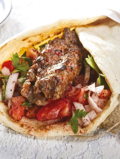Ein frisch zubereitetes Wrap mit saftigem Fleisch, Tomaten, Zwiebeln und Kräutern. Daneben sieht man eine kleine Schale mit weiteren Beilagen. | © Canva