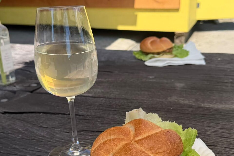Ein köstlicher Burger mit frischem Salat und Tomaten auf einem Tisch, daneben ein Glas Weißwein. Im Hintergrund ist ein gelbes Food-Truck zu sehen. | © Imbissstand_Marhofmüller (c) Schriebl