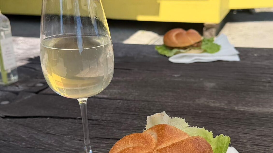 Ein köstlicher Burger mit frischem Salat und Tomaten auf einem Tisch, daneben ein Glas Weißwein. Im Hintergrund ist ein gelbes Food-Truck zu sehen. | © Imbissstand_Marhofmüller (c) Schriebl