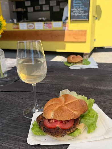 Ein köstlicher Burger mit frischem Salat und Tomaten auf einem Tisch, daneben ein Glas Weißwein. Im Hintergrund ist ein gelbes Food-Truck zu sehen. | © Imbissstand_Marhofmüller (c) Schriebl