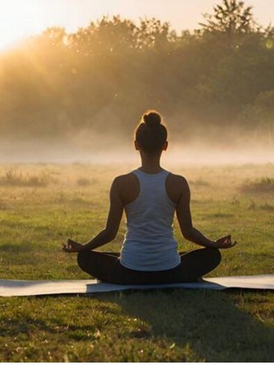 Eine Frau sitzt in den Morgenstunden im Schneidersitz auf einer Yogamatte inmitten einer Wiese. Die Sonne scheint durch die Bäume und der Morgennebel ist deutlich zu sehen. | © Tracky C.