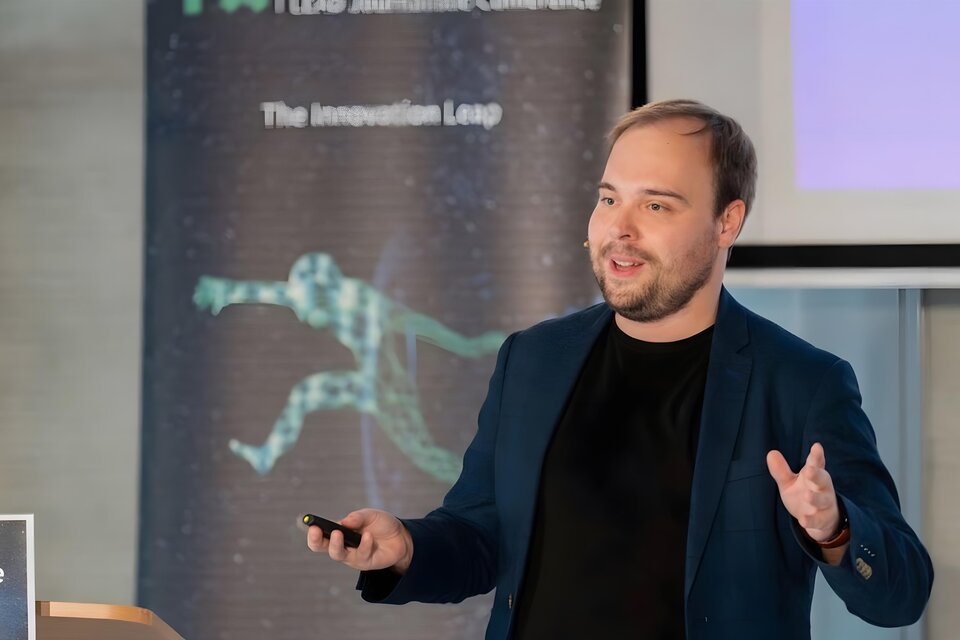 Ein Mann präsentiert mit einem Mikrofon in der Hand. Im Hintergrund sind Grafiken und das Thema "Innovation" zu sehen. | © Görög