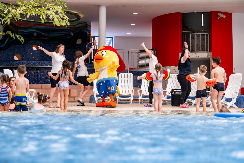 Eine fröhliche Poolparty mit Kindern und einem Maskottchen. Die Kinder genießen das Wasser und tanzen. | © H2O-Hoteltherme GmbH / moving Stills