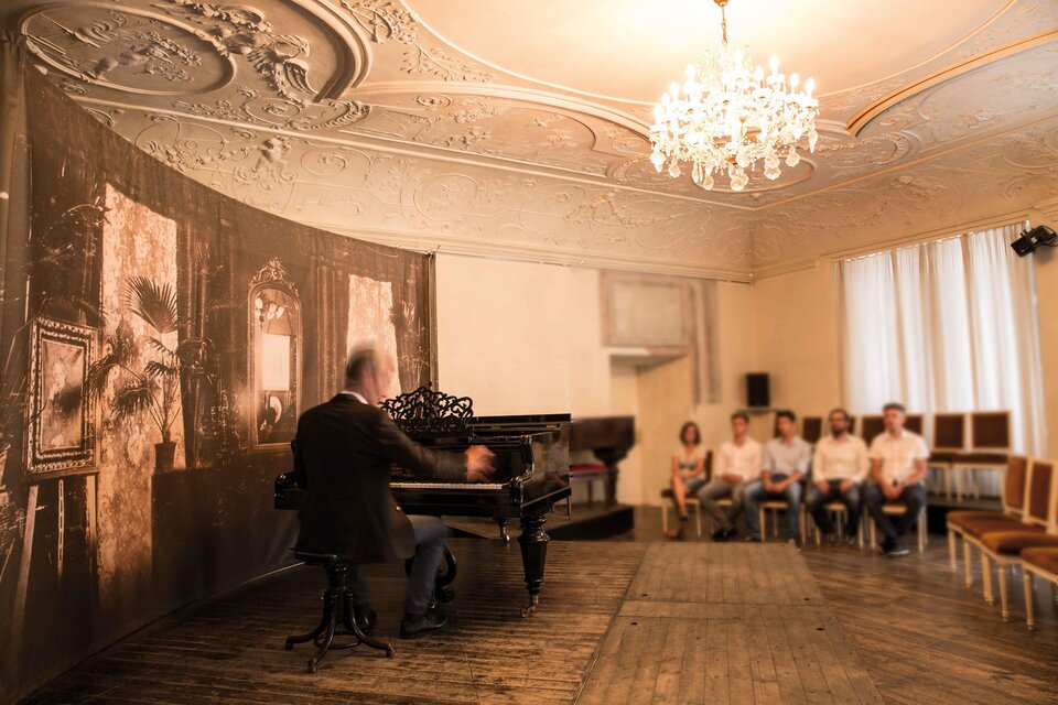 Ein Pianist spielt an einem Klavier in einem eleganten Raum. Eine kleine Gruppe von Zuhörern sitzt aufmerksam und beobachtet das Konzert. | © Königshofer