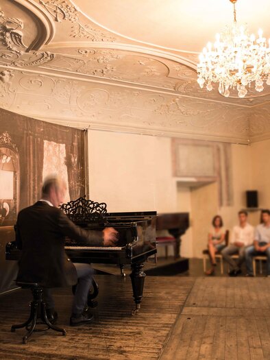 Ein Pianist spielt an einem Klavier in einem eleganten Raum. Eine kleine Gruppe von Zuhörern sitzt aufmerksam und beobachtet das Konzert. | © Königshofer