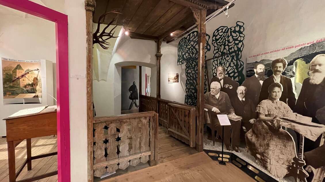 Ein interessanter Ausstellungsraum mit Holzdetails und Fotografien an der Wand. Im Hintergrund sind historische Figuren abgebildet, die die Atmosphäre des Raumes prägen. | © Brahms Museum Mürzzuschlag, Ronald Fuchs