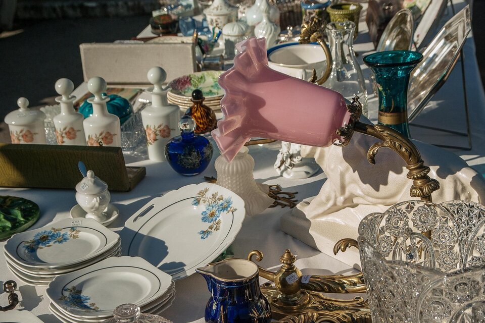 Ein Flohmarkt- oder Antiquitätenstand mit verschiedenen Porzellan-, Glas- und Metallgegenständen. Im Mittelpunkt steht eine auffällige rosa Tischlampe mit verziertem Messingfuß, umgeben von Tellern, Kristallschalen, Vasen und dekorativen Haushaltsgegenständen. | © Pixabay