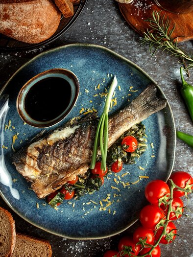 Ein gegrillter Fisch auf einem blauen Teller, serviert mit Kirschtomaten und frischen Kräutern. Im Hintergrund sind einige Brotsorten und grüne Chilis zu sehen. | © Garten Hotel Ochensberger