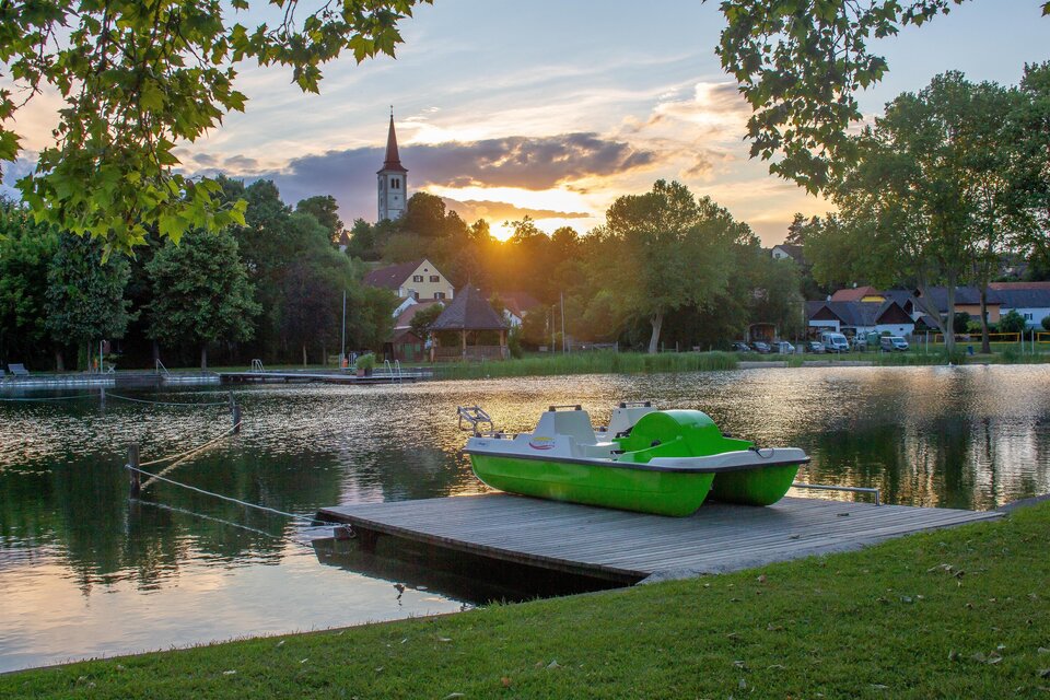 Ein ruhiger See bei Sonnenuntergang mit einem grünen Tretboot auf einem Holzsteg. Im Hintergrund sind Bäume und ein Kirchturm zu sehen. | © Sandra Brünner