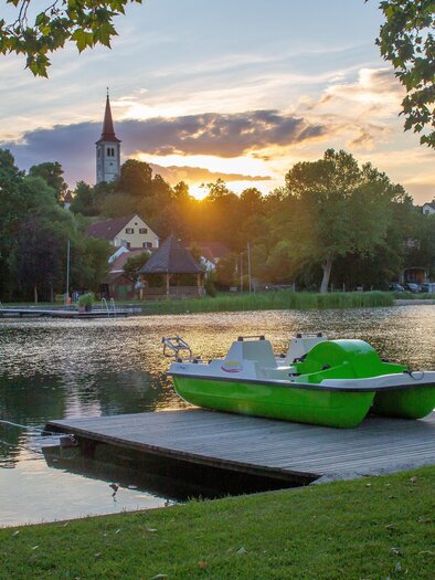 Ein ruhiger See bei Sonnenuntergang mit einem grünen Tretboot auf einem Holzsteg. Im Hintergrund sind Bäume und ein Kirchturm zu sehen. | © Sandra Brünner