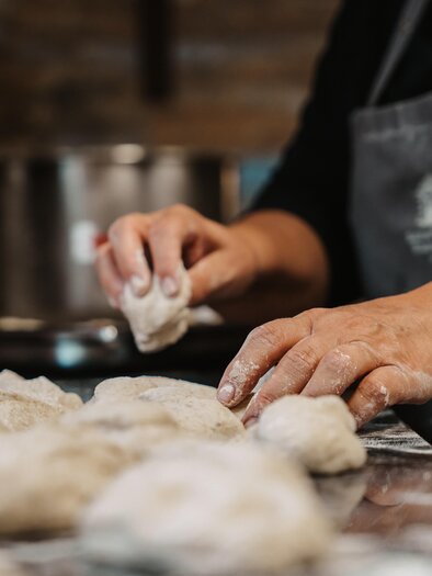 Eine Hand formt Teigstücke auf einer bemehlten Arbeitsfläche. Im Hintergrund sind Kochutensilien zu sehen. | © Michaela Lorber