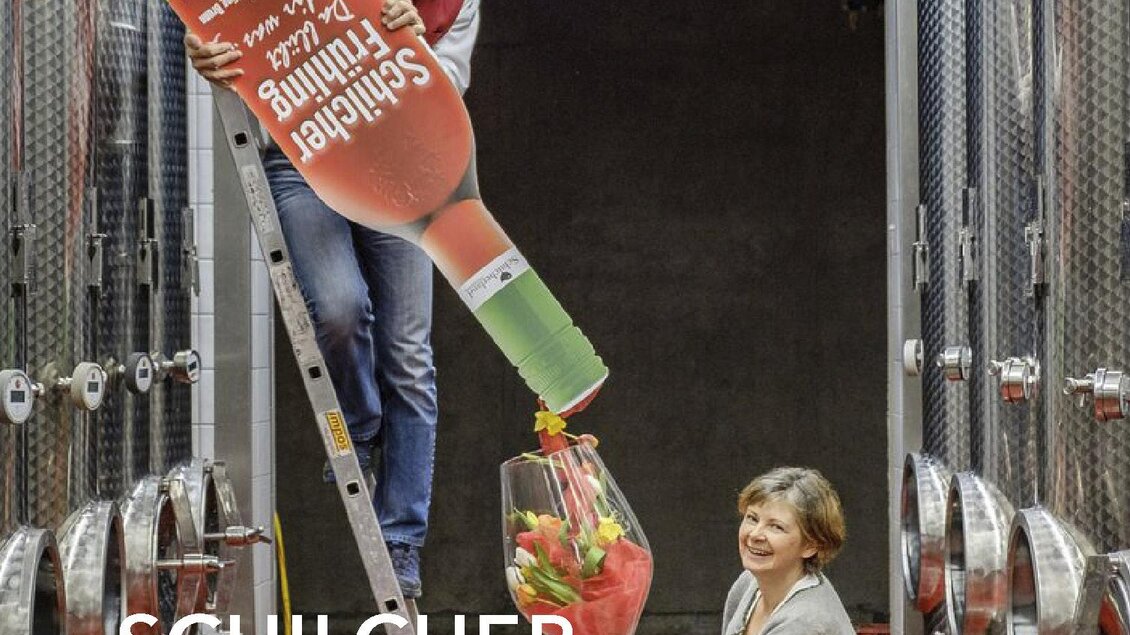 Ein Mann steht auf einer Leiter und gießt eine große Flasche Sekt in ein Glas. Eine Frau steht daneben und hält das Glas, während sie in einer Weinfarm sind. | © @jöbstl