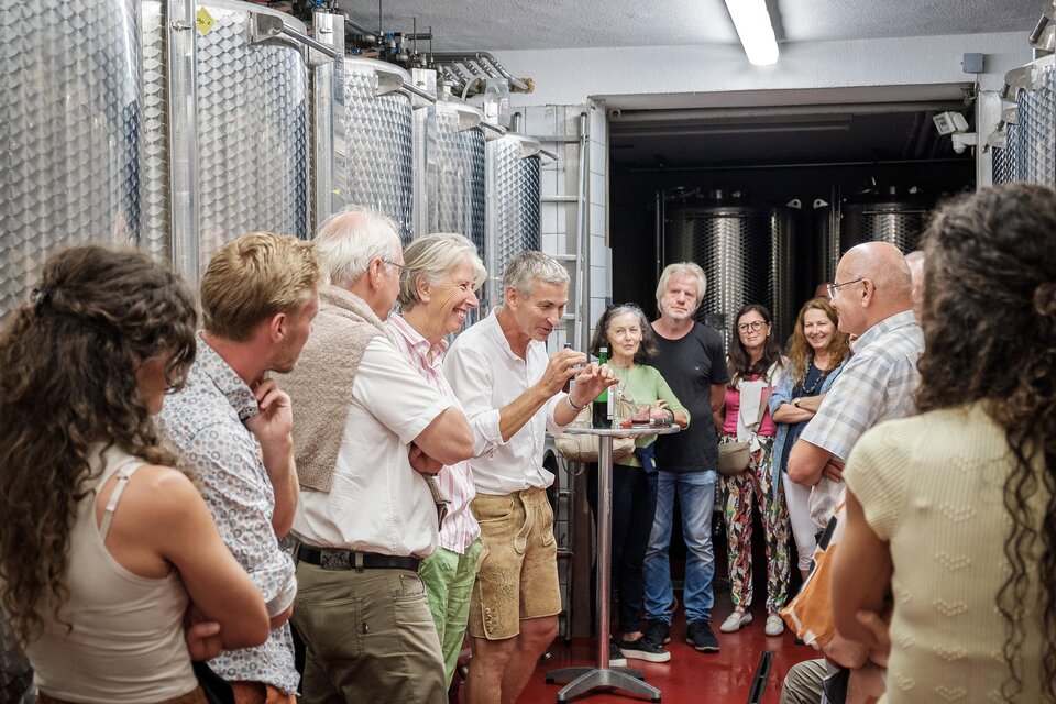 Eine Gruppe von Menschen steht in einer Weinkelterei und hört einem Mann aufmerksam zu. Im Hintergrund sind große Edelstahltanks sichtbar. | © @Jöbst WEingut