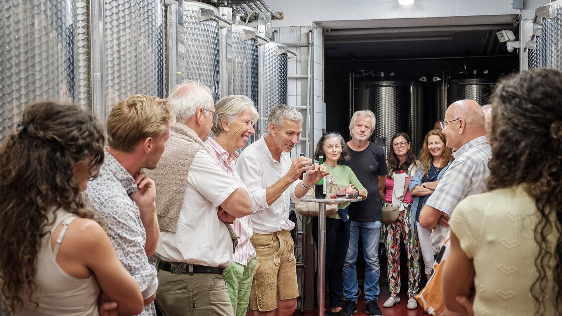 Eine Gruppe von Menschen steht in einer Weinkelterei und hört einem Mann aufmerksam zu. Im Hintergrund sind große Edelstahltanks sichtbar. | © @Jöbst WEingut