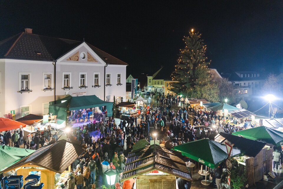 Ein festlicher Weihnachtsmarkt bei Nacht mit zahlreichen Ständen und Menschen. Im Hintergrund steht ein großer, geschmückter Baum.