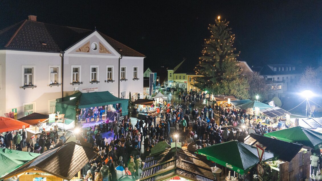 Ein festlicher Weihnachtsmarkt bei Nacht mit zahlreichen Ständen und Menschen. Im Hintergrund steht ein großer, geschmückter Baum.
