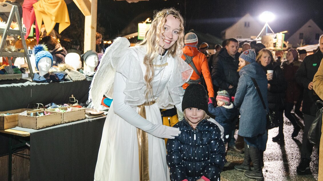 Ein Engel steht lächelnd neben einem Kind auf einem festlichen Markt. Im Hintergrund sind Menschen und Stände mit weihnachtlichen Waren zu sehen.