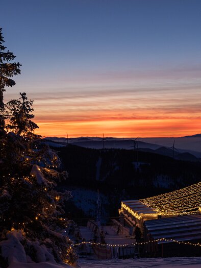 Ein winterlicher Sonnenuntergang über schneebedeckten Hügeln. Ein beleuchteter Baum steht im Vordergrund und ein gemütliches Haus ist im Hintergrund sichtbar. | © nicoleseiser.at