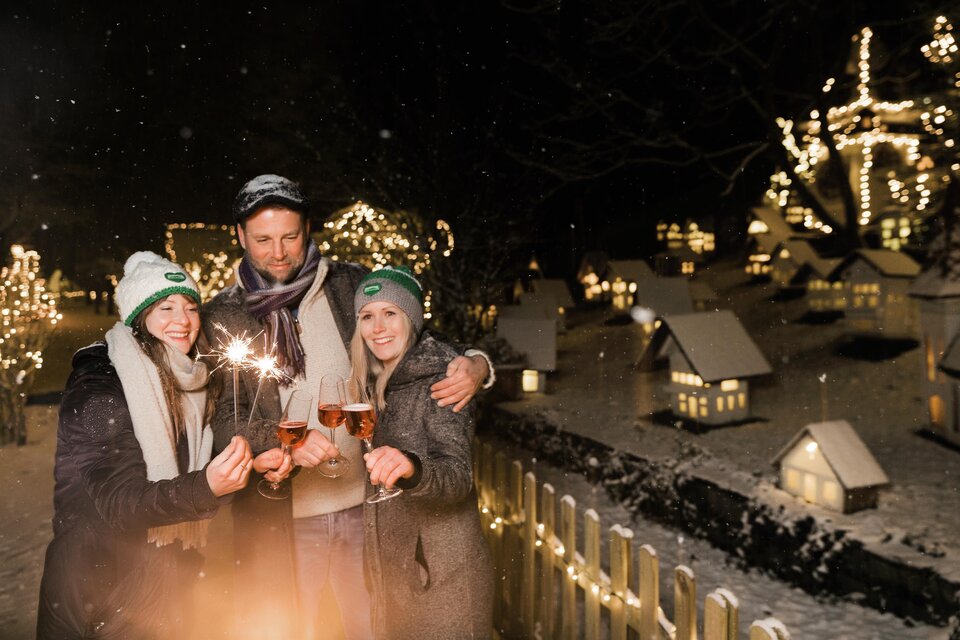 Drei Personen feiern in einer festlich dekorierten Winterlandschaft. Sie halten Sektgläser und Wunderkerzen, umgeben von schneebedeckten Häusern und Lichtern. | © Lupi Spuma