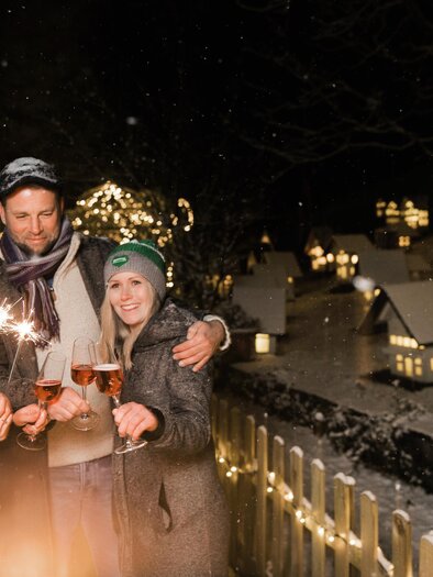 Three people are celebrating in a festively decorated winter landscape. They are holding champagne glasses and sparklers, surrounded by snow-covered houses and lights. | © Lupi Spuma