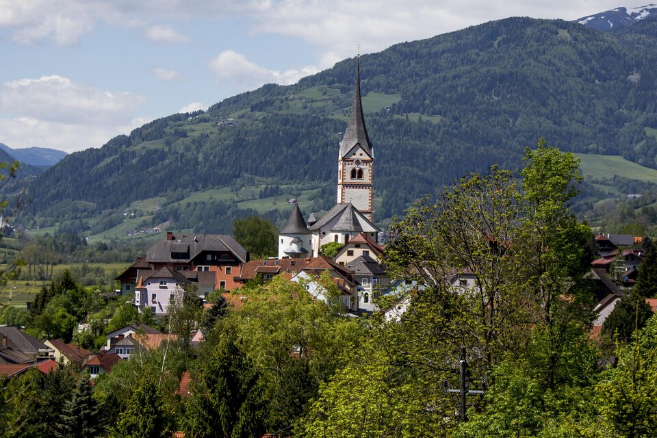 Ein malerisches Dorf mit einer Kirche und einem hohen Kirchturm. Im Hintergrund sind grüne Berge und ein klarer Himmel zu sehen.