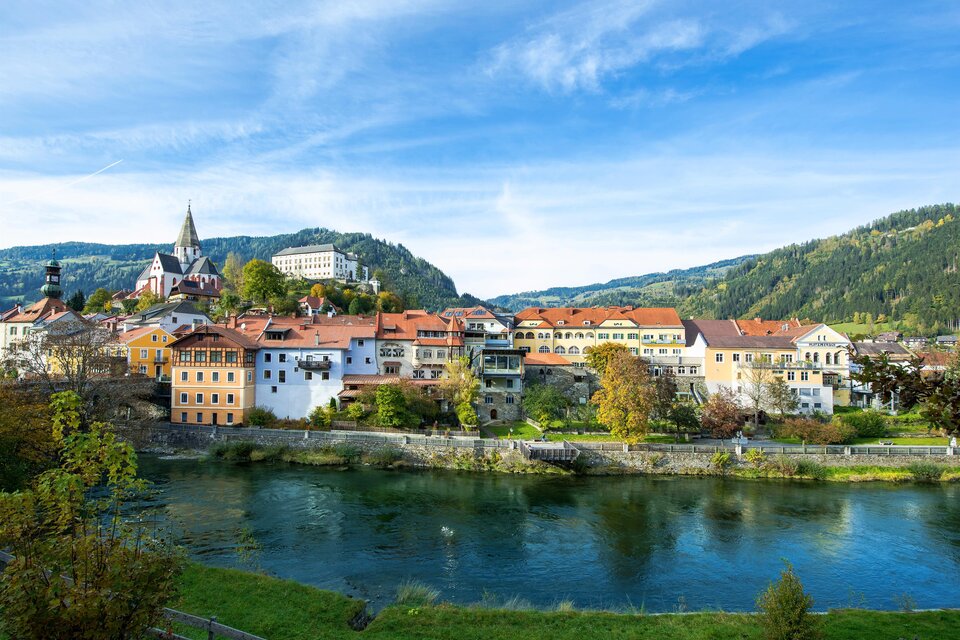 Eine malerische Stadt am Fluss mit bunten Häusern und sanften Hügeln im Hintergrund. Der Himmel ist klar und die Landschaft ist grün.