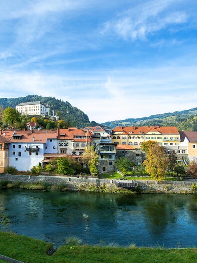 Eine malerische Stadt am Fluss mit bunten Häusern und sanften Hügeln im Hintergrund. Der Himmel ist klar und die Landschaft ist grün.
