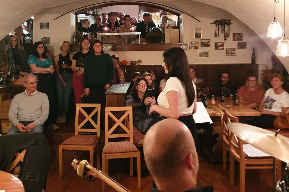 Eine lebendige Veranstaltung in einem gemütlichen Raum mit vielen Zuschauer*innen. Eine Person spricht oder präsentiert im Vordergrund, während die Gäste aufmerksam zuhören. | © Sabine Hermann