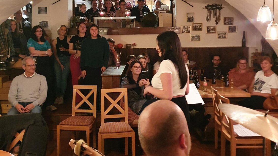 Eine lebendige Veranstaltung in einem gemütlichen Raum mit vielen Zuschauer*innen. Eine Person spricht oder präsentiert im Vordergrund, während die Gäste aufmerksam zuhören. | © Sabine Hermann