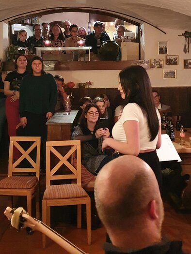 Eine lebendige Veranstaltung in einem gemütlichen Raum mit vielen Zuschauer*innen. Eine Person spricht oder präsentiert im Vordergrund, während die Gäste aufmerksam zuhören. | © Sabine Hermann