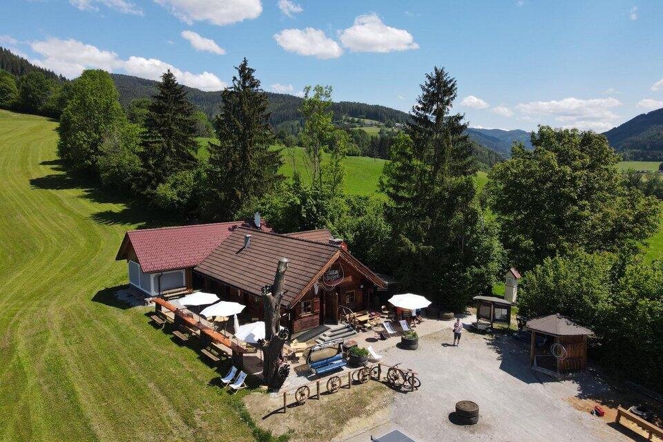Eine idyllische Berghütte umgeben von grüner Wiese und Bäumen. Auf der Terrasse stehen Tische und Stühle unter Sonnenschirmen. | © Marcus Auer