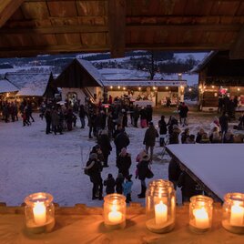 Ein festlicher Weihnachtsmarkt bei Nacht, beleuchtet von Kerzen und Lichtern. Menschen schlendern zwischen den Ständen und genießen die gemütliche Atmosphäre. | © Klaudia Riebenbauer