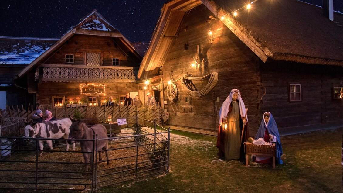 Eine festlich beleuchtete Szene mit traditionellen Holzhäusern und einer Krippe. Dazu stehen zwei Figuren, die die Geburt Jesu darstellen, und einige Tiere sind zu sehen. | © Tourismus Vorau
