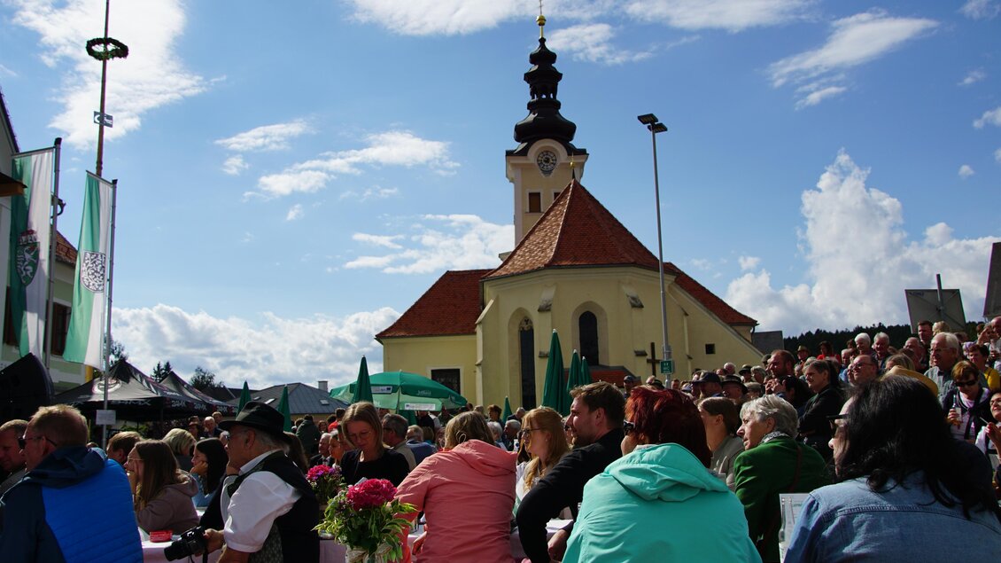 Zu sehen sind Besucher sitzend auf Bänken am Veranstaltungsgelände mit Kirche im Hintergrund. | © Oststeiermark Tourismus