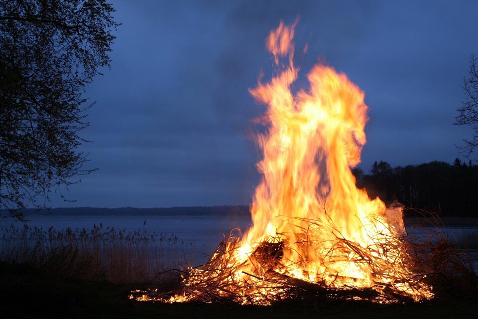 Ein großes Feuer brennt am Ufer eines Sees. Der Himmel ist dunkel und bewölkt.