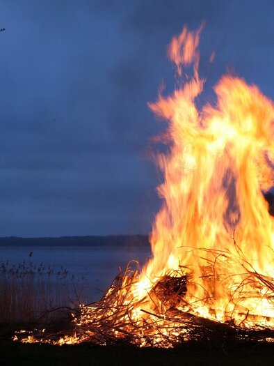 Ein großes Feuer brennt am Ufer eines Sees. Der Himmel ist dunkel und bewölkt.