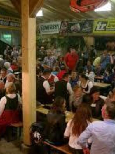 Ein lebhaftes Festival mit vielen Menschen, die an langen Tischen sitzen und feiern. Die Gäste tragen traditionelle Kleidung und die Atmosphäre ist festlich.