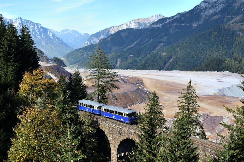 Ein blaublühender Zug fährt über eine historische Brücke inmitten einer malerischen Berglandschaft. Im Hintergrund sind hohe Berge und ein weites, schottriges Tal zu sehen. | © Erzbergbahn