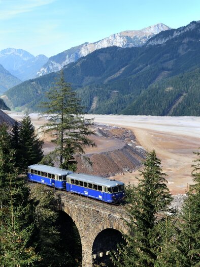 Ein blaublühender Zug fährt über eine historische Brücke inmitten einer malerischen Berglandschaft. Im Hintergrund sind hohe Berge und ein weites, schottriges Tal zu sehen. | © Erzbergbahn