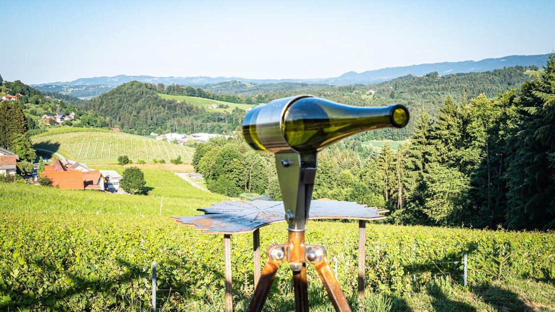 Ein Weinfass auf einem Stand inmitten von Weinbergen. Im Hintergrund erstrecken sich Hügel und Bäume unter einem klaren Himmel. | © @weingut Jöbstl