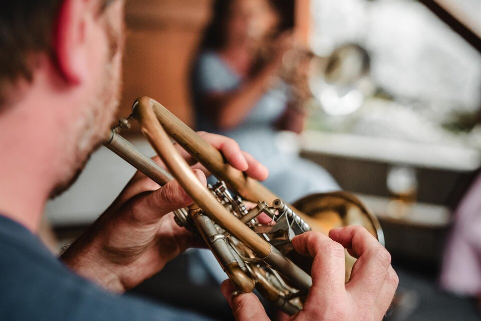Ein Musiker spielt Trompete in einer gemütlichen Umgebung. Im Hintergrund ist eine weitere Person zu sehen, die ebenfalls ein Instrument spielt.