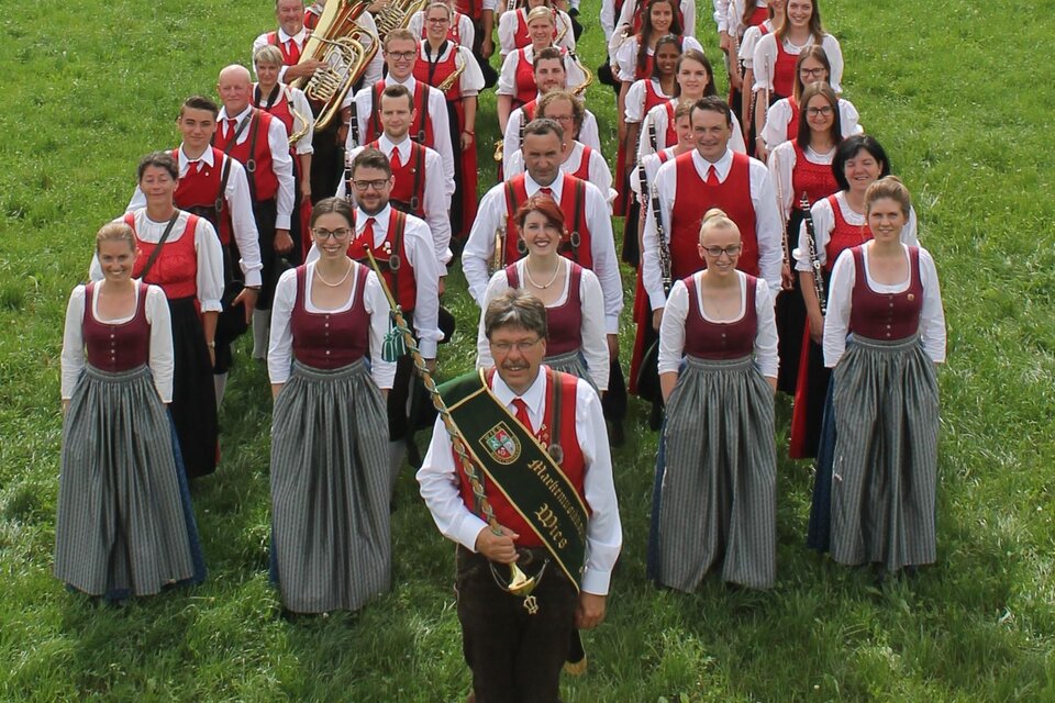 Eine Gruppe von Menschen in traditionellen Trachten steht auf einer grünen Wiese. Einige halten Musikinstrumente und lächeln in die Kamera. | © MMK Wies