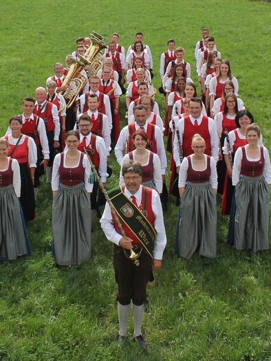 Eine Gruppe von Menschen in traditionellen Trachten steht auf einer grünen Wiese. Einige halten Musikinstrumente und lächeln in die Kamera. | © MMK Wies