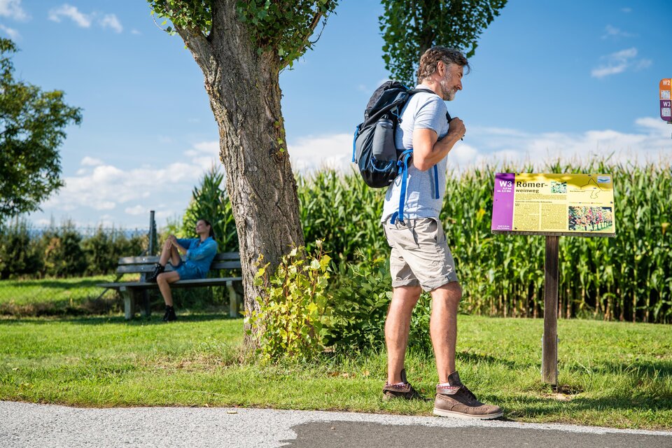 Ein Mann mit einem Rucksack steht vor einem Hinweisschild in der Natur. Im Hintergrund sitzt eine andere Person auf einer Bank. | © Mias Photoart
