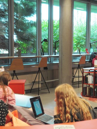 In einer modernen Bibliothek arbeiten mehrere Personen an Laptops. Große Fenster lassen viel Tageslicht herein und schaffen eine angenehme Atmosphäre. | © M. Ohrt